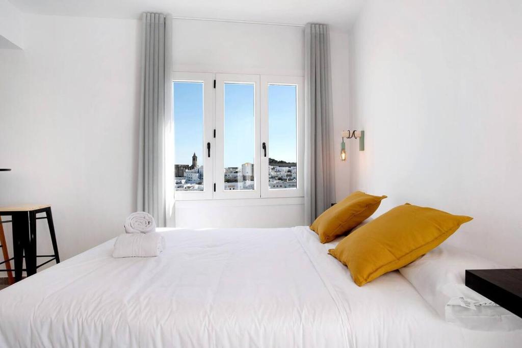 Cama blanca con almohadas amarillas y ventana en Apartamentos Vexèr, en Vejer de la Frontera