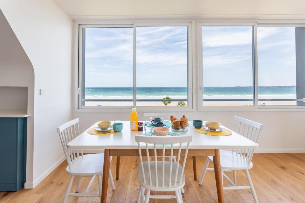 uma sala de jantar branca com uma mesa e cadeiras e o oceano em La Grande Plage em Saint-Cast-le-Guildo