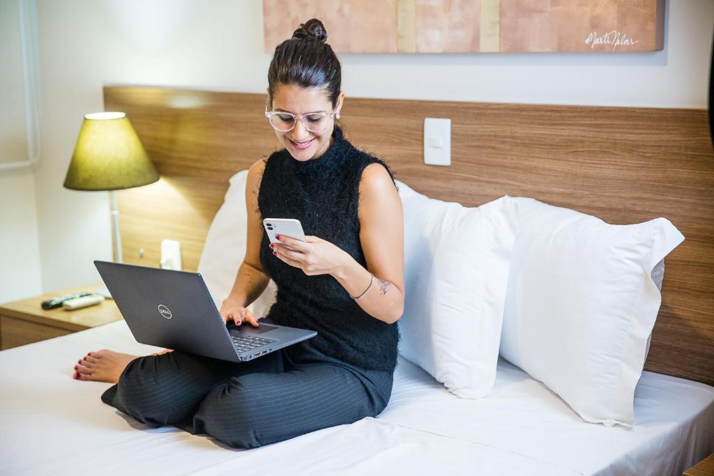 a woman sitting on a bed with a laptop and a phone at Ville Hotel Gramadão in Votuporanga