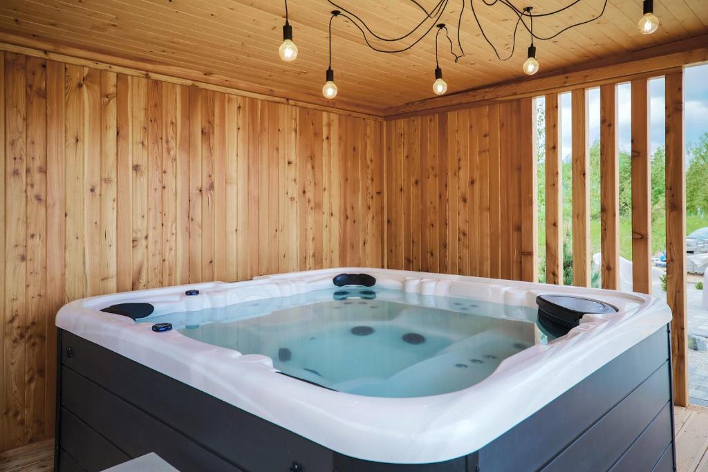 Large white hot tub inside wooden pavilion with hanging Edison bulbs and slatted outdoor view.