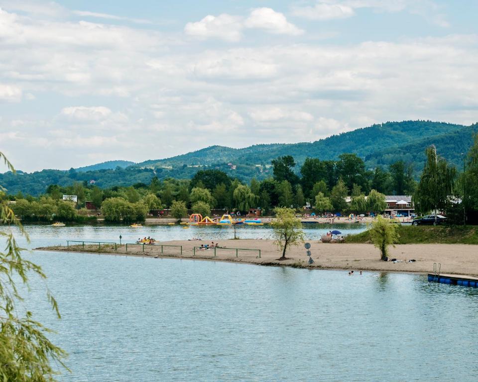Calm lake shoreline with sandy beach, distant water inflatables, and forested hills beyond.