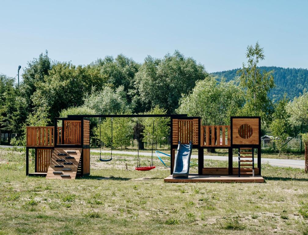 Wooden playground structure with swings and slide on grassy field against tree-lined backdrop.