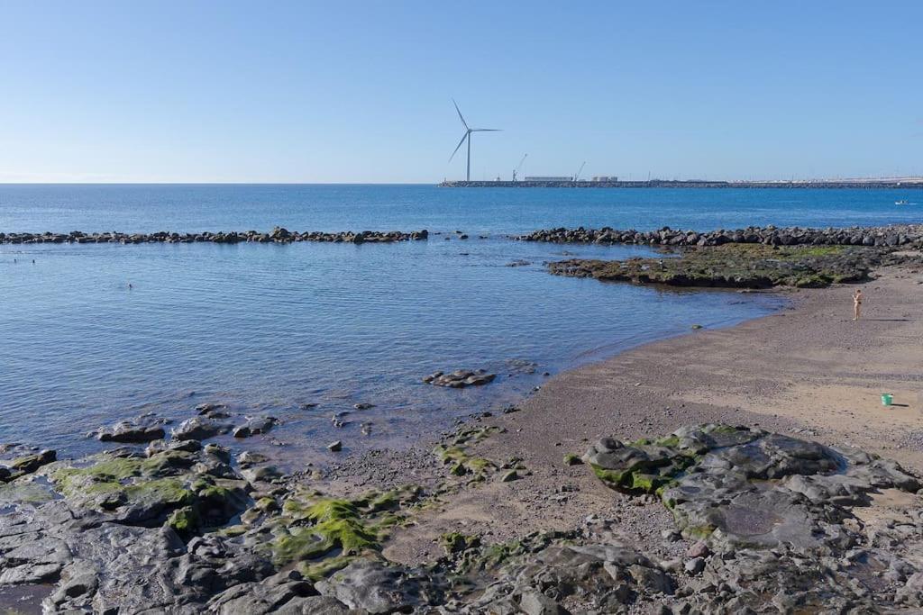 a beach with rocks and water and a windmill in the distance at Las Afortunadas , Playa Arinaga 1ºA in Arinaga