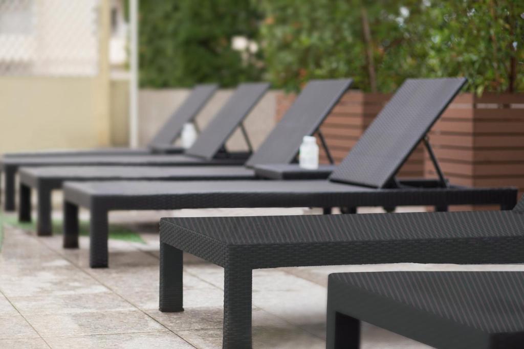 A row of dark woven sun loungers on a paved pool deck beside planted screening.
