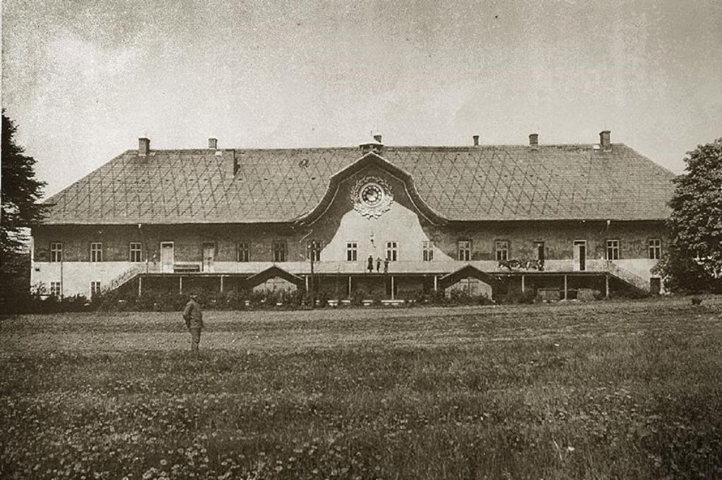 Sepia-toned historic manor with ornate central gable, grassy foreground and lone figure walking