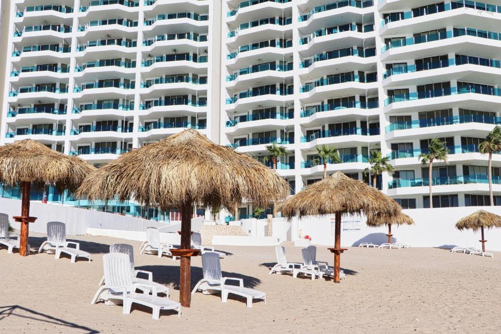 a beach with chairs and straw umbrellas and a large building at Esmeralda Beach Resort in Puerto Peñasco