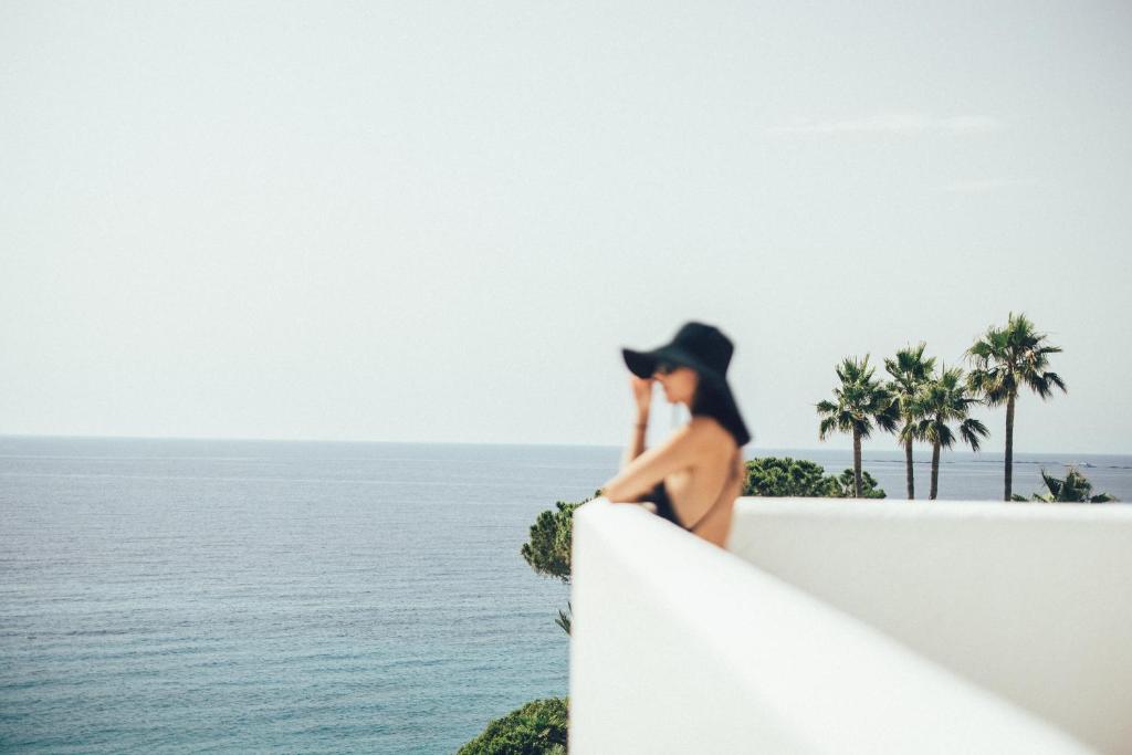 a woman in a hat sitting on a wall looking at the ocean at Cala Lanuza Suites in El Campello