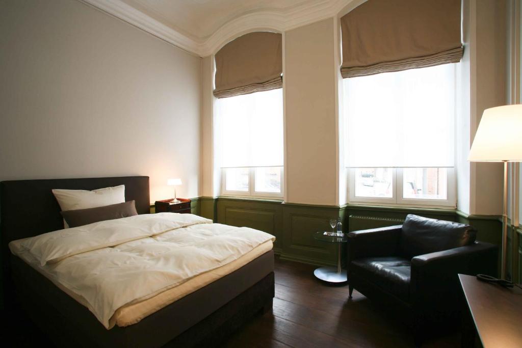 a bedroom with a bed and a chair and two windows at Hotel Anno 1216 in Lübeck