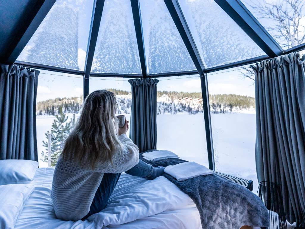 Kobieta siedząca na łóżku patrząca przez okno w obiekcie Lake Igloo Ukkohalla w mieście Hyrynsalmi