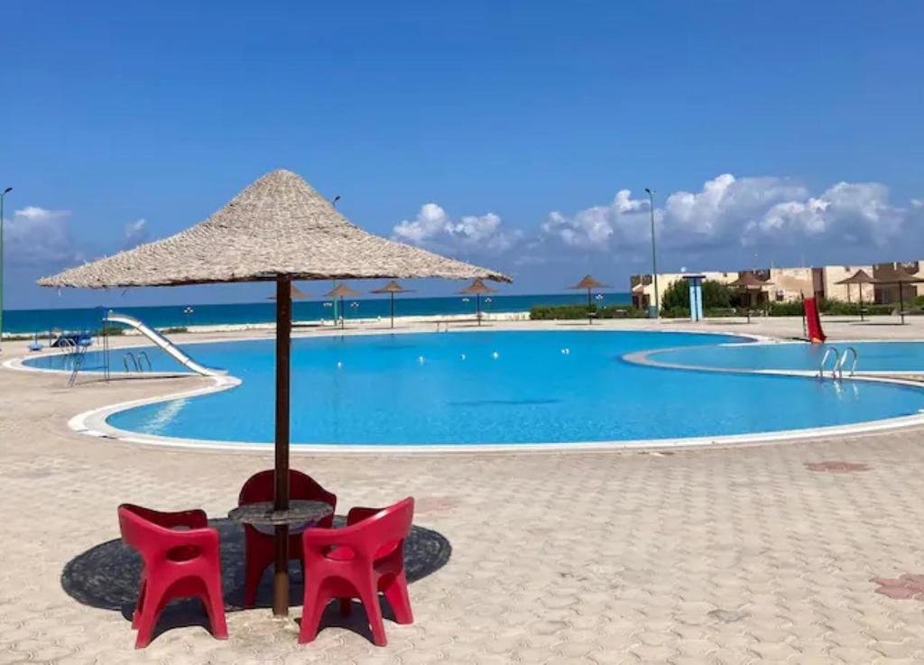 una mesa, dos sillas y una sombrilla frente a una piscina en Location Maison en bord de mer, Nord Egypte en Al Ḩammām