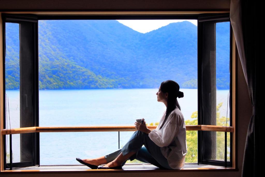 Yutorelo Nikko في نيكو: امرأة جالسة على حافة النافذة تطل على بحيرة