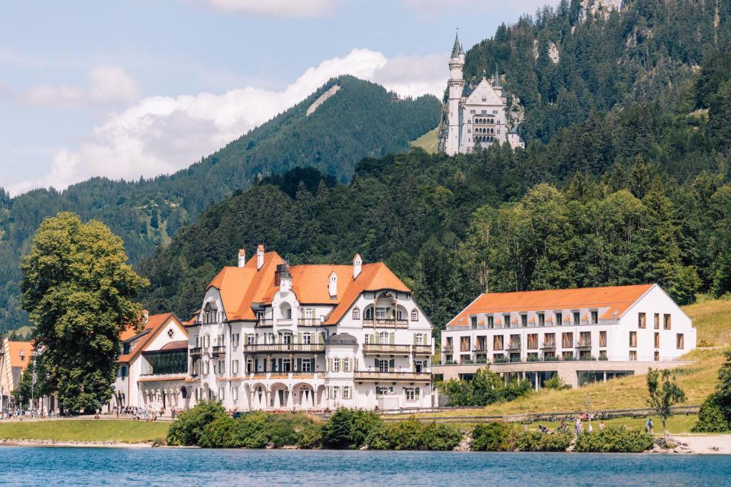 duży budynek nad brzegiem jeziora z zamkiem w obiekcie AMERON Neuschwanstein Alpsee Resort & Spa w mieście Schwangau