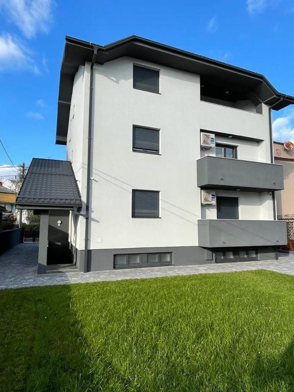 a white building with a black roof and green grass at Apartmani Daisy in Velika Gorica