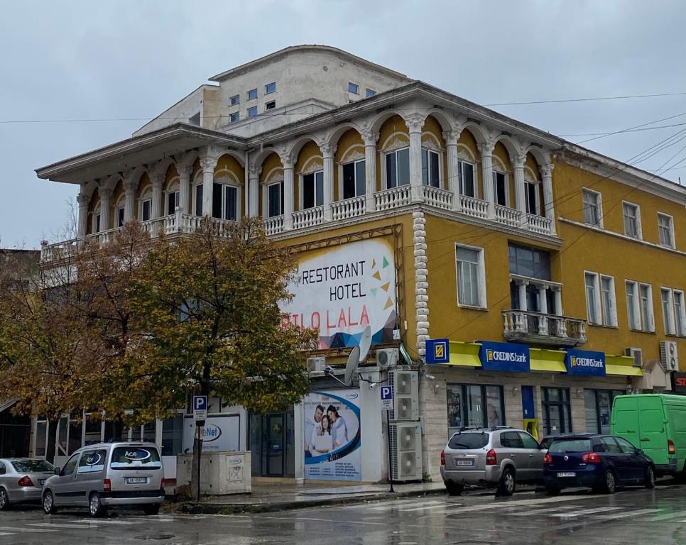 un bâtiment jaune dans une rue avec des voitures garées devant dans l'établissement Pilo Lala Hotel, à Lushnjë
