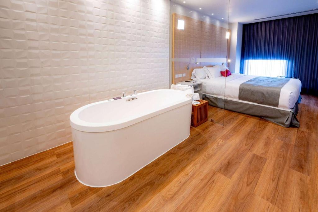 a bathroom with a bath tub in front of a bed at Ramada Encore by Wyndham Guadalajara Aeropuerto in Guadalajara