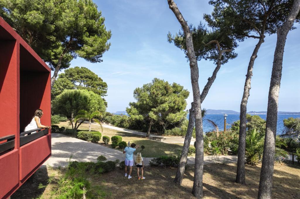 two children standing on the balcony of a red house with trees at Belambra Clubs Presqu&#39;île De Giens - les Criques in Hyères