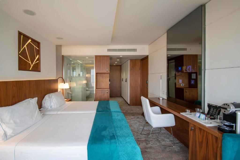 A spacious-looking room with a double bed, seating, minibar area, and glass bathroom entrance.