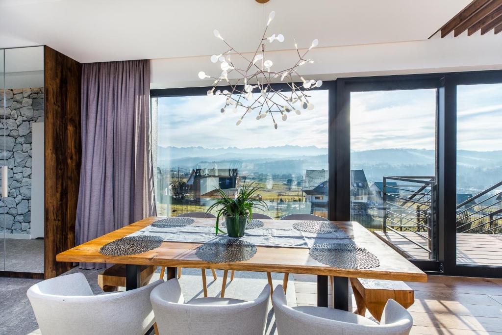 Sunlit dining area with wooden table, modern chandelier and floor-to-ceiling windows overlooking landscape.