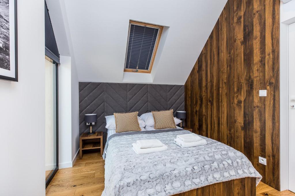 Well-lit bedroom with plush grey bedding, bedside lamps, skylight above and warm wooden flooring.