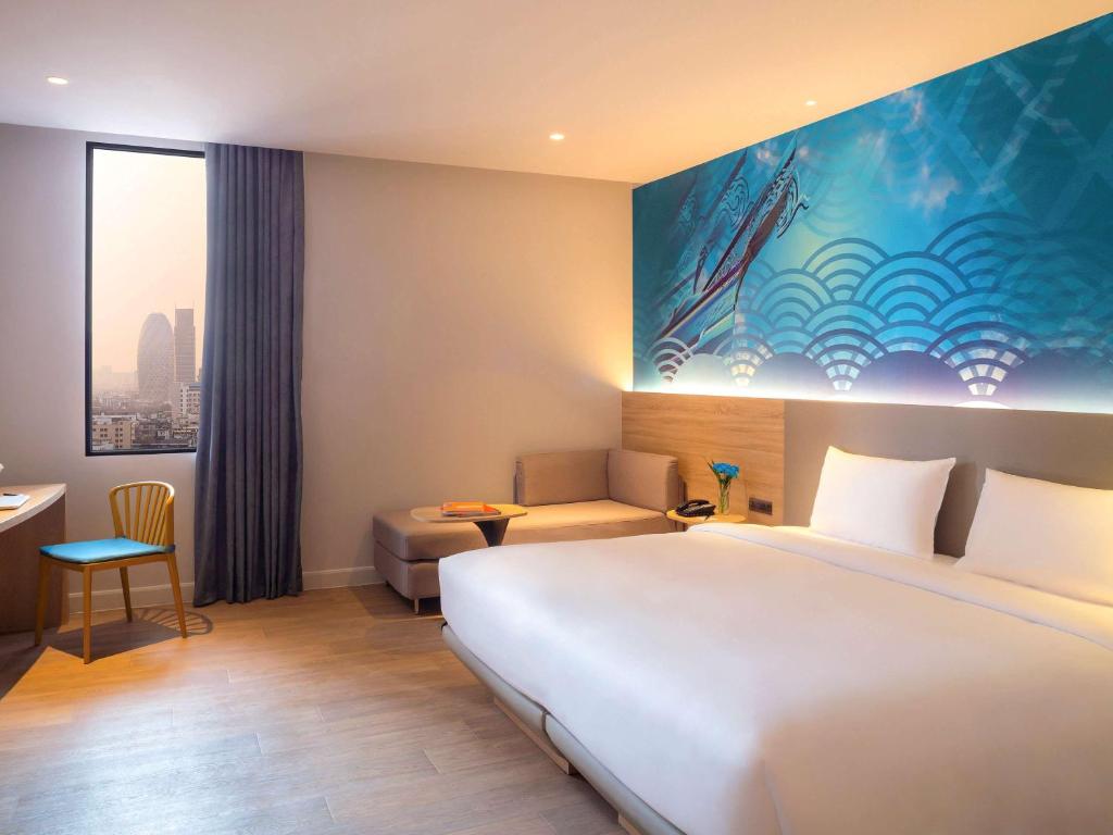 A bright guest room with a large white bed, sofa, desk, chair and a narrow city-facing window.