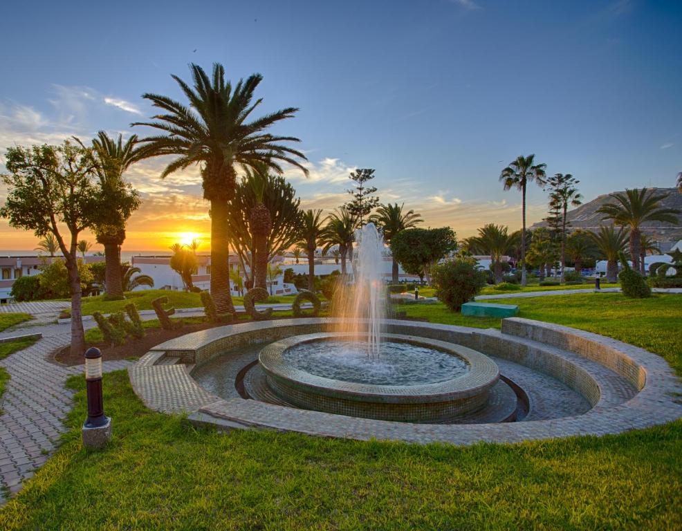 Hotel Club Almoggar Garden Beach في أغادير: وجود نافورة في حديقة مع غروب الشمس في الخلفية