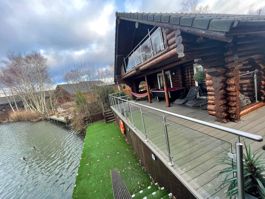 a house with a deck next to a body of water at Woodpecker Log Cabin with hot tub, pizza oven bbq entertainment area, lakeside with private fishing peg situated at Tattershall Lakes in Tattershall