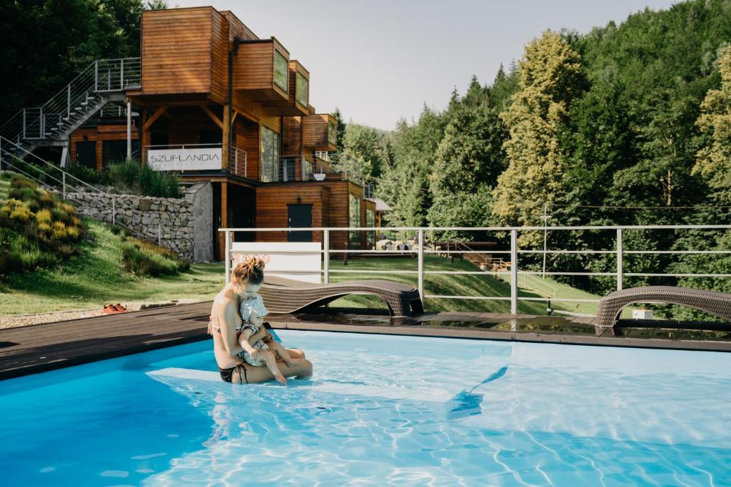 Woman holding a child while sitting in an outdoor pool, modern wooden cabins and forested backdrop.