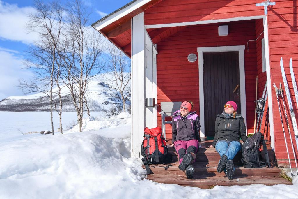 dos personas sentadas fuera de una casa roja en la nieve en Kilpisjärven Retkeilykeskus Cottages, en Kilpisjärvi