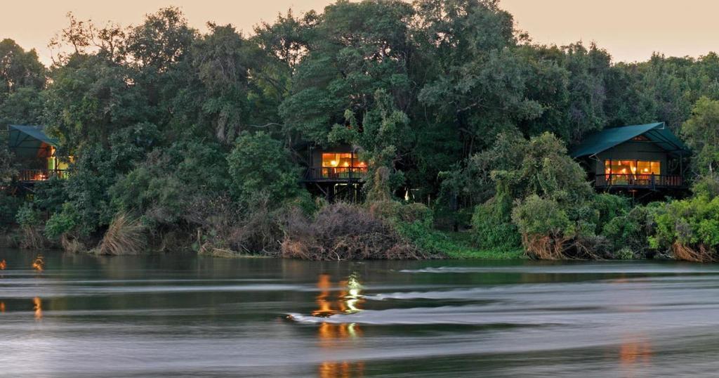 a couple of houses on a river with trees at Tintswalo Siankaba in Livingstone