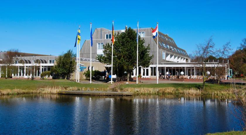 un gran edificio con banderas frente a un lago en Fijn App HELIOS, eigen KEUKEN op beg grond met Prive TERRAS, Incl Verwarmd Binnen-ZWEMBAD, Nabij Duinen Strand en Vuurtoren, en Hollum