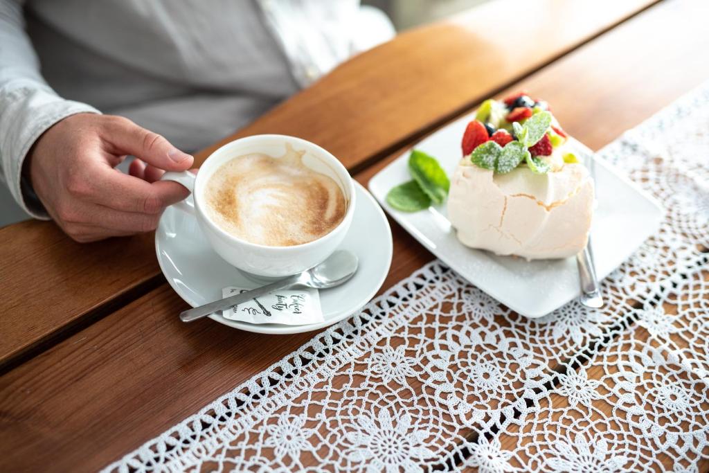 Hand holding cappuccino beside a pavlova dessert on a lace-covered wooden table