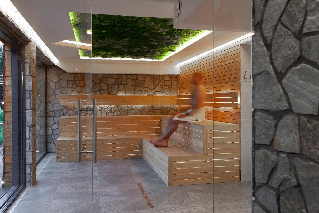 Blurred person sitting on wooden sauna bench beneath a green moss ceiling, stone walls and glass door.