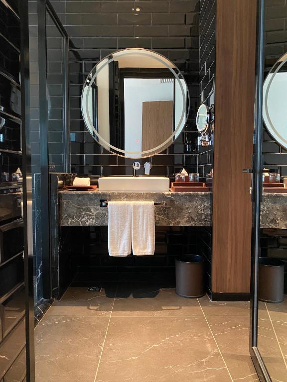 A dark-tiled bathroom vanity with a round illuminated mirror and marble-effect counter.