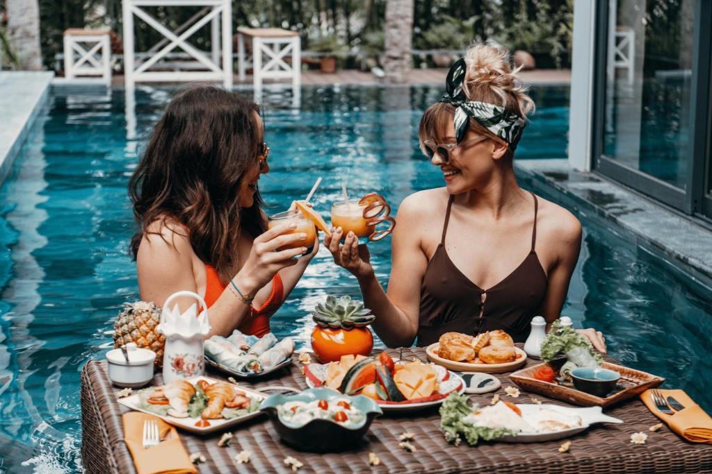 dwie kobiety siedzące przy stole z jedzeniem obok basenu w obiekcie G Boutique Resort Hoi An w Hoi An