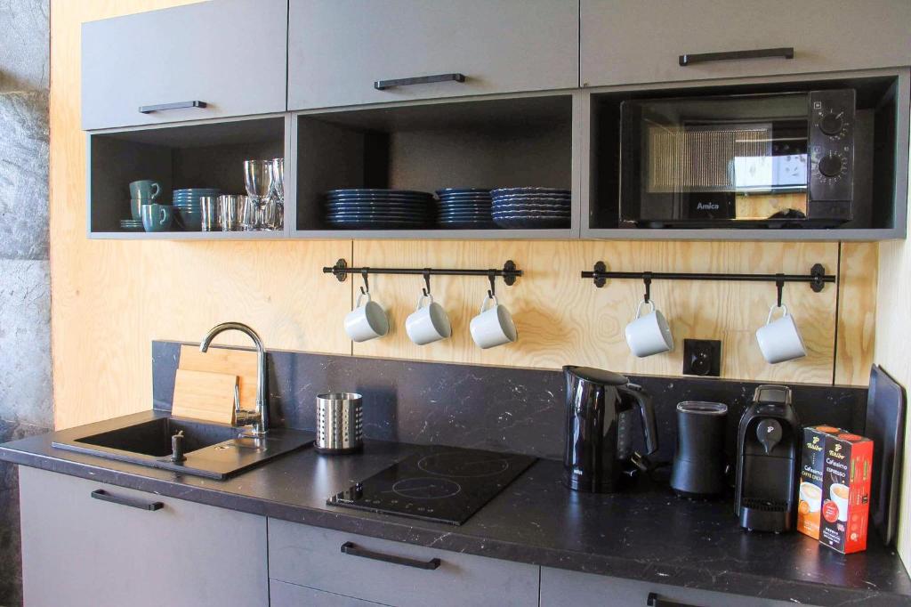 Compact modern kitchenette with black countertops, hanging mugs, microwave, kettle, coffee machine, and open shelving.
