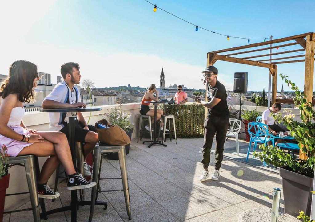 a group of people sitting on a rooftop with a man standing on a patio at Hostel Vertigo Nîmes Gare Centre in Nîmes