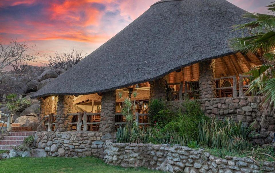 a log cabin with a thatched roof and a stone wall at Ondundu Lodge in Kamanjab