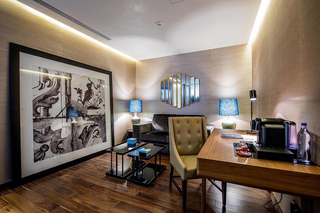 A compact hotel workspace with a wooden desk, armchair, artwork, and drinks station.