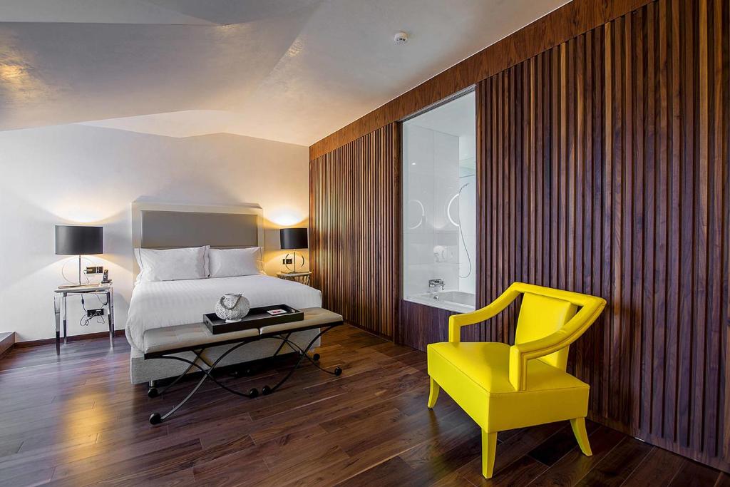 A spacious-looking bedroom with a bright yellow accent chair and a glass-sided bathroom zone.