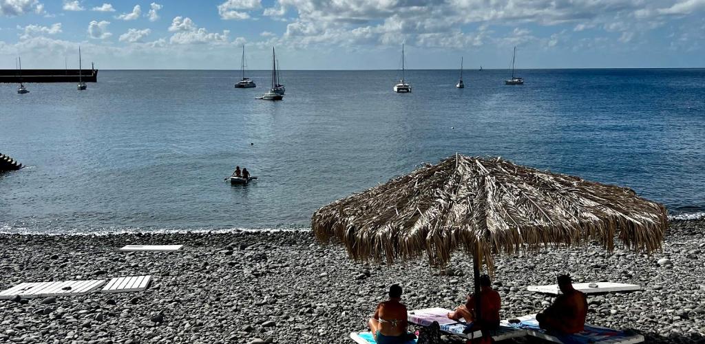 grupa ludzi siedzących pod słomianym parasolem na plaży w obiekcie Home Away From Home w mieście Caniçal