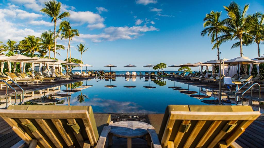 a pool with chairs and umbrellas and the ocean at Four Seasons Resort Hualalai in Kaupulehu