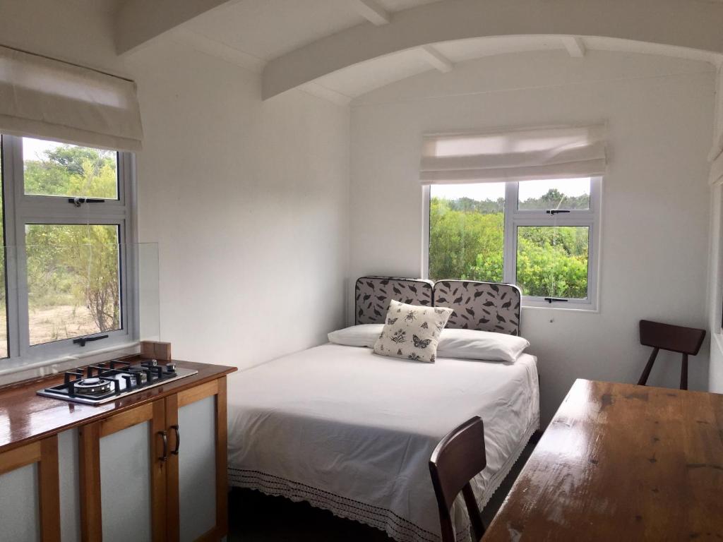 a bedroom with a bed and a desk and two windows at Fynbos Tiny Home - Off Grid, Surrounded by Nature in Plettenberg Bay