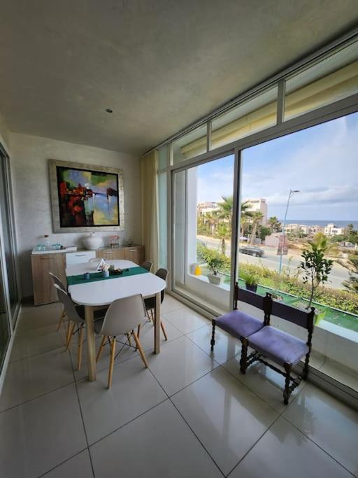 une salle à manger avec une table, des chaises et une grande fenêtre dans l'établissement Melrose Beach - Apartment with a beach view, à Dar Bouazza
