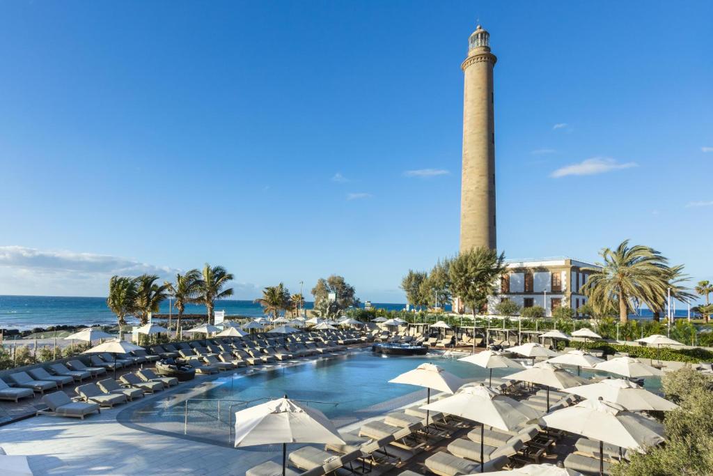 a pool with umbrellas and a lighthouse in the background at Hotel Faro, a Lopesan Collection Hotel - Adults Only in Maspalomas