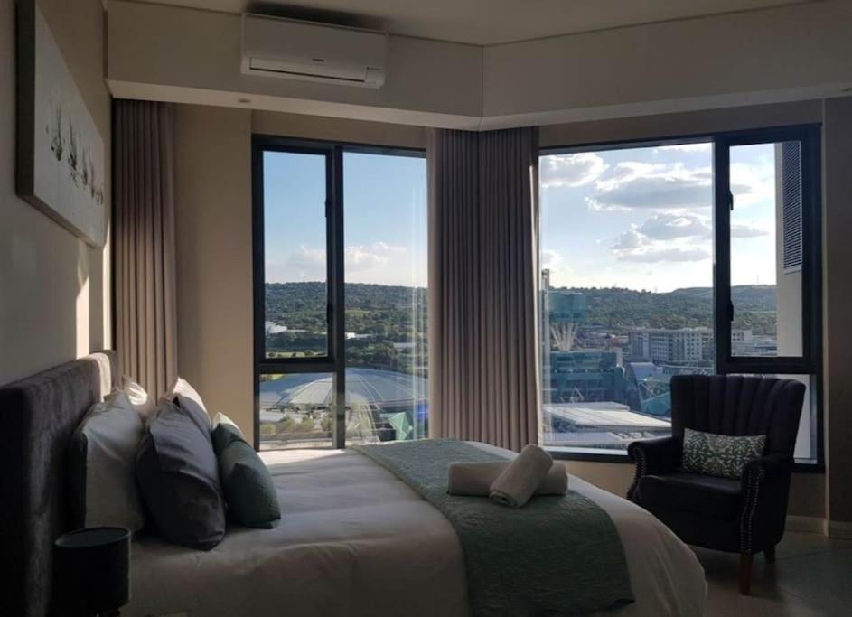 a bedroom with a bed and a chair and large windows at Luxury 1 bedroom apartment in vibrant Menlyn Maine in Pretoria