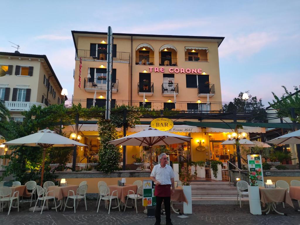 a man standing in front of a building with tables and umbrellas at Hotel Tre Corone in Garda