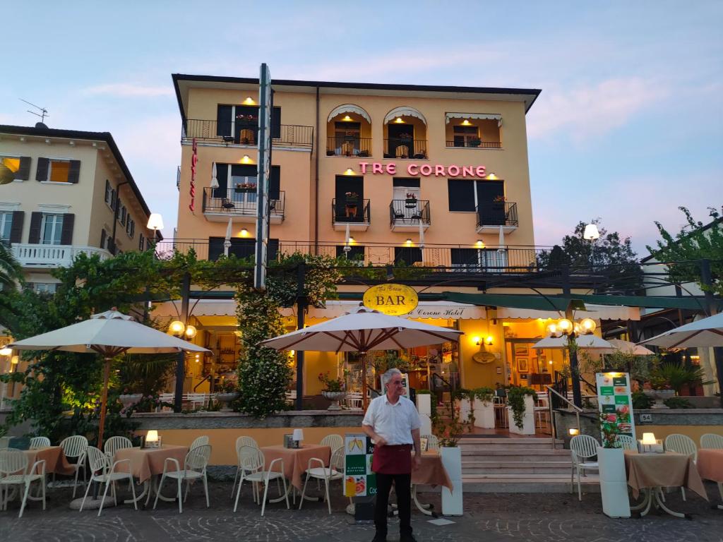 Ein Mann steht vor einem Gebäude mit Tischen und Sonnenschirmen in der Unterkunft Hotel Tre Corone in Garda