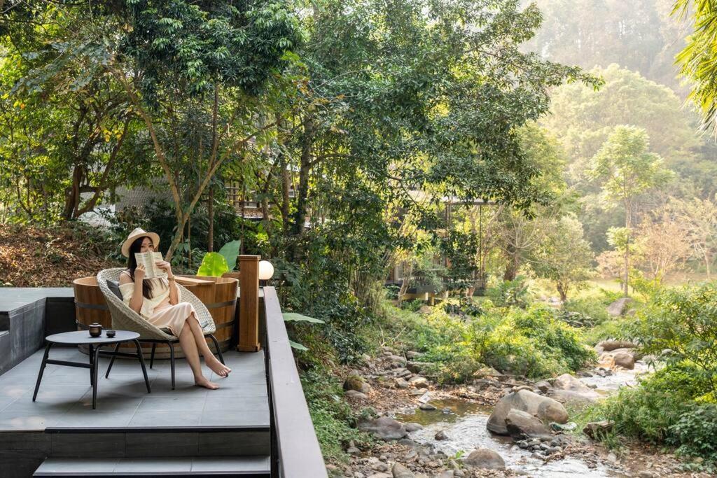 a woman sitting in a chair reading a book on a wooden deck at Zen MaeKampong Waterfall Villa in Ban Huai Kaeo