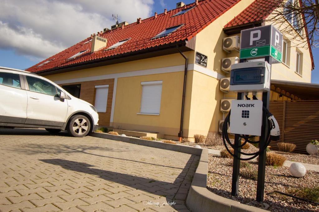 White car parked beside a Noatk electric vehicle charging station outside a yellow house with red roof.