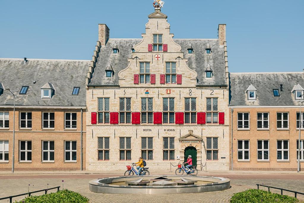 zwei Personen, die vor einem großen Gebäude Fahrrad fahren in der Unterkunft Hotel St Joris I Kloeg Collection in Middelburg